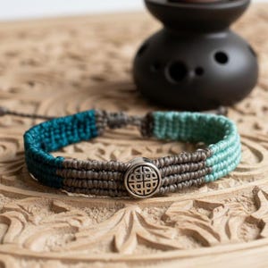 Puede incluir: Una pulsera de macramé con un dije circular plateado en el centro que presenta un patrón de nudo geométrico. La banda está hecha de cordón tejido en tres secciones distintas: azul verdoso profundo, gris neutro y verde menta claro.