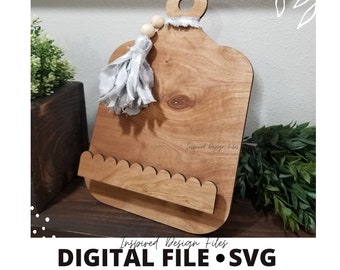 Könnte beinhalten: Ein Holzbrett zum Schneiden mit einem gewellten Rand und einem kleinen Regal. Das Brett hat eine hellbraune Farbe und einen weißen Quasten, der von oben hängt. Der Text "Inspired Design Files" ist auf das Brett geschrieben.