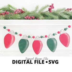Op de afbeelding: Een kerstkrans met rode en groene lampvormen die aan elkaar geregen zijn met witte en zilveren kralen. De krans hangt aan een witte schouw. De tekst "Inspired Design Files DIGITAL FILE. SVG" staat onderaan de afbeelding.