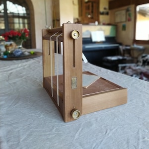 Può includere: Un telaio in legno con due pomelli in ottone e una corda bianca che lo attraversa. Il telaio è appoggiato su una tovaglia bianca. Il testo "TOGHETHER" è visibile sul telaio.