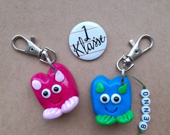 Könnte beinhalten: Zwei bunte Monster-Schlüsselanhänger mit Glitzer, einer pink und einer blau, mit einem silbernen Schlüsselring und einem Knopf mit der Aufschrift "1 Klasse".