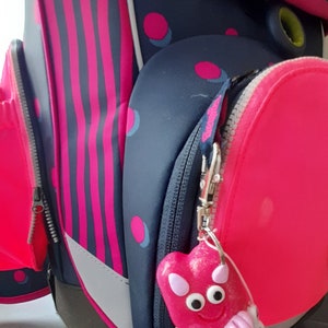 K&ouml;nnte beinhalten: Nahaufnahme eines Schulrucksacks in Rosa und Marineblau. Der Rucksack hat einen herzf&ouml;rmigen Anh&auml;nger in Rosa mit einem l&auml;chelnden Gesicht und dem Namen "ALE" an einer Schnur. Der Rucksack hat rosa Streifen und Punkte.