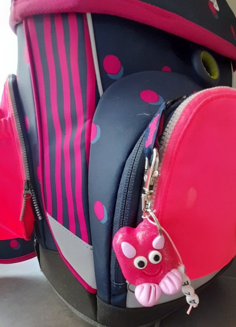 K&ouml;nnte beinhalten: Nahaufnahme eines Schulrucksacks in Rosa und Marineblau. Der Rucksack hat einen herzf&ouml;rmigen Anh&auml;nger in Rosa mit einem l&auml;chelnden Gesicht und dem Namen "ALE" an einer Schnur. Der Rucksack hat rosa Streifen und Punkte.