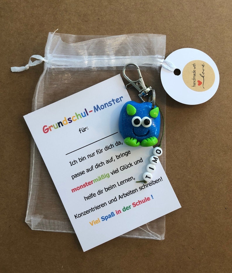 K&ouml;nnte beinhalten: Ein blauer Monster-Schl&uuml;sselanh&auml;nger mit gr&uuml;nen Akzenten und dem Namen "TIMO" auf Perlen. Der Schl&uuml;sselanh&auml;nger ist an einer Karte mit deutschem Text befestigt, einschlie&szlig;lich "Grundschul-Monster" und einem "handmade with love"-Etikett.