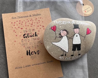 Könnte beinhalten: Handbemalter Flussstein mit Braut und Bräutigam, die herzförmige Ballons halten, mit den Namen "Theresia & Steffen" und dem Datum "10.05.2025". Enthält eine Kraftpapierkarte und einen kleinen Geschenkebeutel.