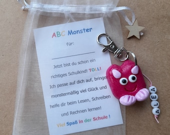 Könnte beinhalten: Ein pinkfarbener Monster-Schlüsselanhänger mit dem Namen "ALEA" an einer Schnur, befestigt an einem silbernen Verschluss. Der Schlüsselanhänger befindet sich neben einer Karte in einem durchsichtigen weißen Beutel mit der Aufschrift "ABC Monster" und Text auf Deutsch.