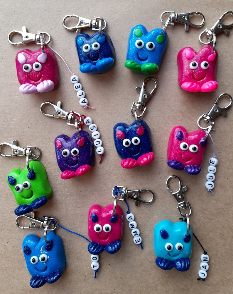 K&ouml;nnte beinhalten: Eine Sammlung handgefertigter Schl&uuml;sselanh&auml;nger. Jeder Schl&uuml;sselanh&auml;nger zeigt ein l&auml;chelndes, abstraktes Wesen in Pink, Blau, Gr&uuml;n und Lila, mit silberfarbenen Verschl&uuml;ssen. Einige Schl&uuml;sselanh&auml;nger haben Namensschilder mit Perlen.