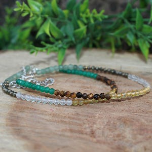 Puede incluir: Tres pulseras de cuentas con una variedad de piedras preciosas de colores. Las pulseras presentan cuentas verdes, marrones, amarillas y transparentes, con detalles de cadena plateada. Las pulseras se exhiben sobre una superficie de madera.