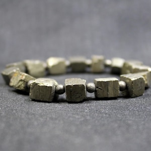 May include: A bracelet made of pyrite cubes, separated by small black beads. The bracelet is made of natural stone and has a rustic, earthy look.