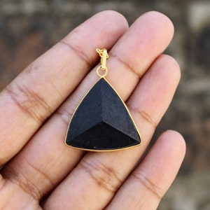 Puede incluir: Un colgante triangular negro con un marco y una argolla dorados. El colgante tiene una superficie facetada, que capta la luz. La joya es probablemente un colgante de collar, adecuado para varios estilos.