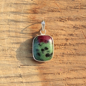 Può includere: Un ciondolo rettangolare con un'ansa argentata. Il ciondolo presenta una grande pietra preziosa lucida con un vivace mix di colori. La parte superiore è di un rosso intenso, che sfuma in una base verde e nera maculata. La pietra è incastonata in una lunetta d'argento.