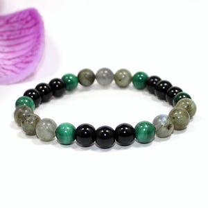 Könnte beinhalten: Ein Armband aus runden Perlen in Schwarz, Grün, Grau und schillernden Farben. Das Armband wird auf einer weißen Oberfläche präsentiert. Im Hintergrund ist ein lila Blütenblatt zu sehen. Die Perlen haben einen Durchmesser von ca. 0,8 cm.