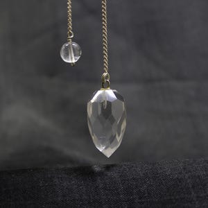 May include: A clear, faceted crystal pendulum with a silver chain and a small, round crystal bead. The pendulum hangs against a dark backdrop, emphasising its geometric form and transparency. The design is simple and elegant.