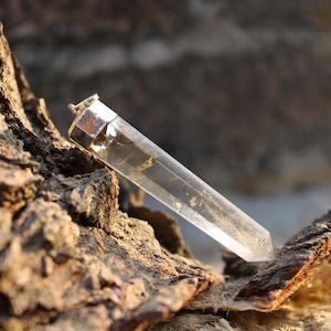 Può includere: Un pendente di cristallo di quarzo trasparente con un cappuccio in metallo color argento. Il cristallo è allungato e sfaccettato, con un'estremità appuntita. Il pendente è esposto su una superficie marrone strutturata.