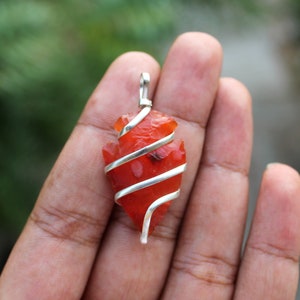 May include: A carnelian arrowhead pendant wrapped in silver wire. The carnelian is a deep orange-red colour and the wire is a shiny silver.