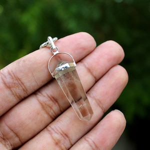 Peut inclure: Un pendentif en cristal de quartz transparent avec un sertissage en argent. Le pendentif est en forme de pointe et est suspendu à une chaîne en argent.