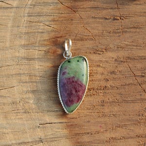 May include: A gemstone pendant with a silver-colored bail. The pendant features a polished, teardrop-shaped stone with a green and burgundy color pattern. The stone is set in a silver-colored bezel with a textured edge. The pendant is displayed on a wooden surface.