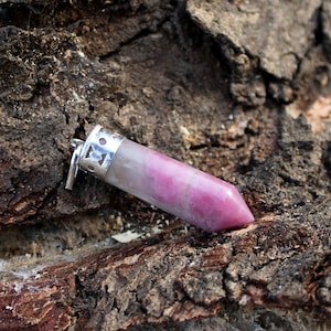 May include: A pink and white crystal pendant with a silver-colored metal cap and bail. The crystal is a polished, elongated hexagonal shape. The pendant is set against a textured, brown bark background.