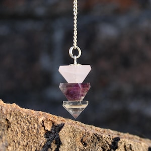 Peut inclure: Une chaîne argentée soutient un pendentif composé de trois formes de cristal facettées. Le cristal supérieur est rose clair, le milieu est violet et le bas est transparent. Le pendentif est présenté sur un fond flou et texturé.