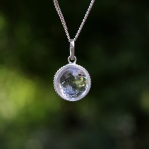 Può includere: Una collana con catena d'argento con un ciondolo di cristallo trasparente. Il ciondolo è rotondo e ha una cornice d'argento.