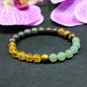 Könnte beinhalten: Ein Armband mit Perlen aus bernsteinfarbenen, grünen und dunkelgrauen Edelsteinen. Die Perlen sind kreisförmig angeordnet. Das Armband wird auf einer dunklen Oberfläche präsentiert. Ein stilvolles Accessoire mit Edelsteinen in verschiedenen Größen und Formen.