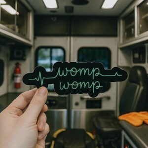May include: Black sticker with the words "womp womp" in a green, stylized font, with a green heartbeat line. The sticker is held in front of an ambulance interior.