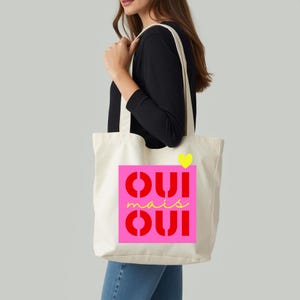 Könnte beinhalten: Cremefarbene Tragetasche mit einem pinkfarbenen Quadrat mit den Worten "OUI mais OUI" in Rot und Gelb. Ein gelbes Herz befindet sich über dem Text. Die Tasche hat lange Träger und wird über der Schulter getragen.