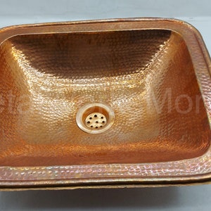 May include: A square copper sink with a hammered finish and a drain in the centre. The sink has a raised edge around the perimeter.