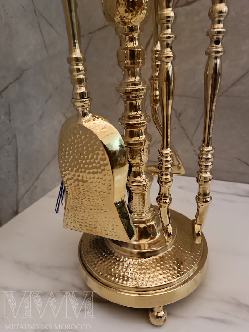 May include: A gold metal fireplace tool set with a hammered finish. The set includes a shovel, poker, and tongs. The tools are mounted on a stand with a round base.