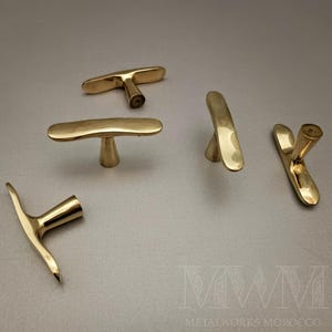 May include: Five brass cabinet pulls in various shapes, including T-bar and curved designs. The hardware has a polished finish and is arranged on a neutral gray surface. The logo "METALWORKS MOROCCO" is visible in the bottom right corner.
