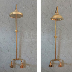 May include: A vintage-style, gold-colored shower fixture. The showerhead is round and flat, with a curved arm extending from the wall. The fixture includes a vertical pipe and two faucet handles at the base. The background is a gray marble-like wall.