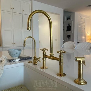May include: A gold kitchen faucet with two handles and a separate sprayer. The faucet has a bridge design and is mounted on a white countertop. The faucet is labeled "MWM METALWORKS MOROCCO".
