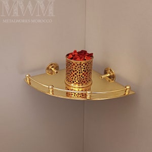 Op de afbeelding: Gouden hoekplank met decoratief metaalwerk en een kleine gouden container gevuld met rode bloemblaadjes. De plank is aan een muur gemonteerd.