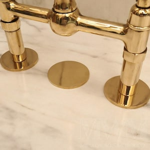 Könnte beinhalten: Eine polierte goldfarbene Brückenarmatur aus Metall mit zwei vertikalen zylindrischen Sockeln und einer zentralen runden Abdeckplatte. Die Armatur hat eine glatte, reflektierende Oberfläche und ist auf einer weißen Marmorplatte montiert.