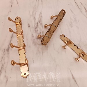May include: Three brass coat racks with a decorative design. One rack has three hooks, while the other two have two hooks each. The racks are displayed on a marble surface.