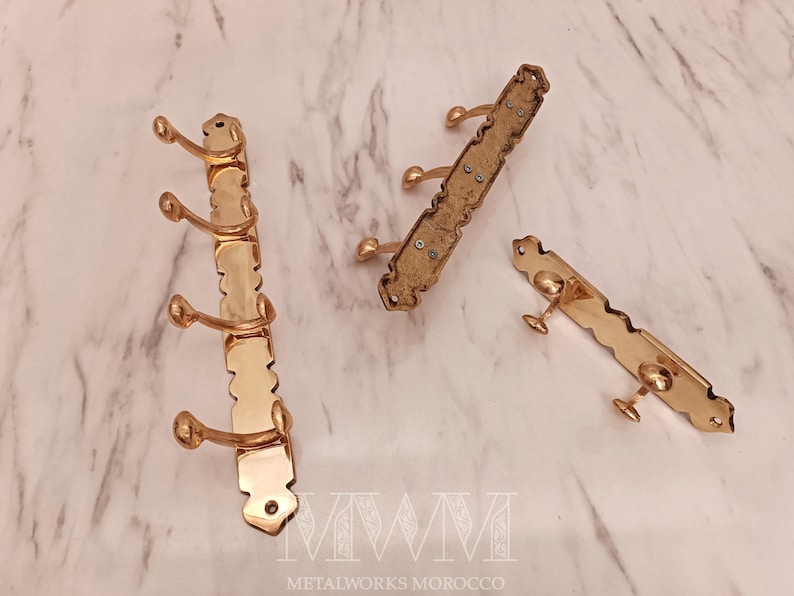 May include: Three brass coat racks with a decorative design. One rack has three hooks, while the other two have two hooks each. The racks are displayed on a marble surface.