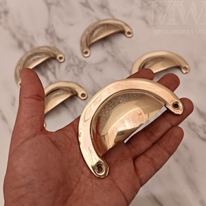 May include: Five brass cabinet knobs with a rounded, curved shape. The knobs have a polished, shiny finish. The knobs are displayed on a marble surface.