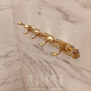 May include: A gold-colored metal wall hook with four curved hooks. The hook is mounted on a rectangular base with decorative detailing. The background is a marble-patterned wall. The text "METALWORKS MOROCCO" is at the bottom.