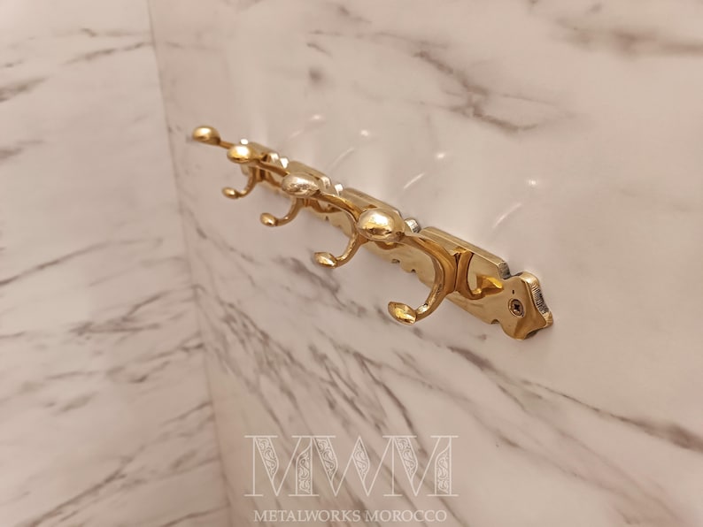 May include: A gold-colored metal wall hook with four curved hooks. The hook is mounted on a rectangular base with decorative detailing. The background is a marble-patterned wall. The text "METALWORKS MOROCCO" is at the bottom.