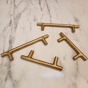May include: Four gold-toned metal cabinet pulls with a hammered texture. The pulls are in the shape of a T with a rounded end.