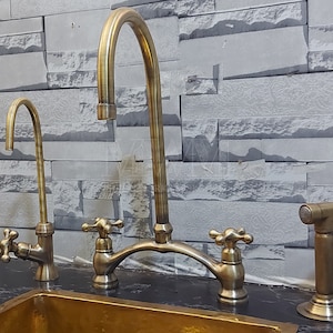 May include: A brass kitchen faucet with a high arc spout and two handles. The faucet has a brushed finish and is mounted on a countertop. There is a separate brass sprayer on the right side of the faucet.