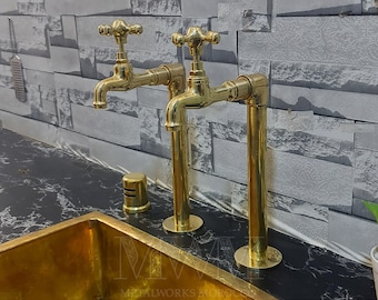 Puede incluir: Dos grifos de lavabo dorados con un diseño vintage, con una manija en forma de cruz y un caño largo y cilíndrico. Los grifos están montados en una encimera negra con un lavabo dorado en primer plano.