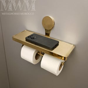 May include: A gold-colored metal toilet paper holder with a shelf, holding two rolls of white toilet paper and a black smartphone. The holder is mounted on a gray wall. The text "METALWORKS MOROCCO" is visible.