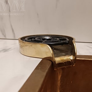 K&ouml;nnte beinhalten: Ein goldfarbenes Waschbecken mit einem schwarzen Metall-Ablaufstopfen. Der Ablaufstopfen hat ein kreisf&ouml;rmiges Design mit einem kleinen Auslauf in der Mitte.