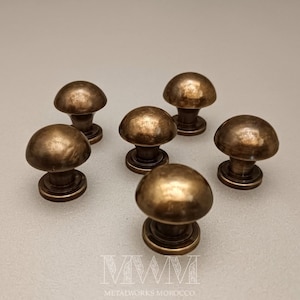 May include: Six bronze mushroom-shaped cabinet knobs. The knobs have a rounded top and a circular base. The metal has a slightly aged appearance. The text "MWM METALWORKS MOROCCO" is at the bottom.