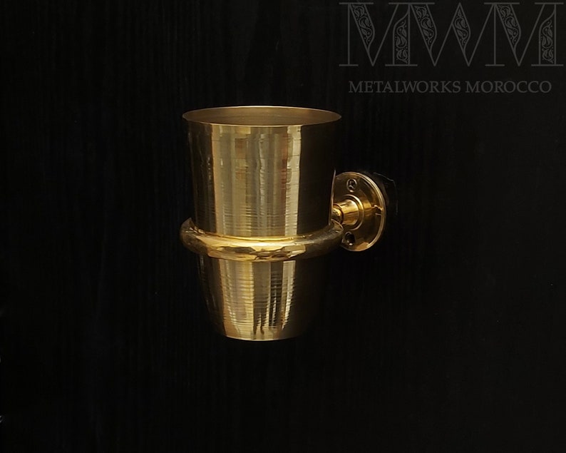 May include: A gold-toned metal wall-mounted toothbrush holder with a hammered finish. The holder is attached to a black wall.