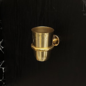 May include: A gold-toned metal toothbrush holder with a round base and a cylindrical cup. The holder is mounted to a black wall.