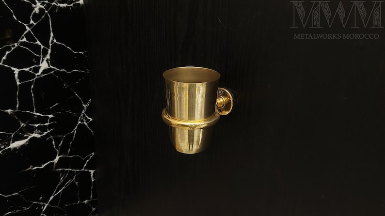 May include: A gold-toned metal toothbrush holder with a round base and a cylindrical cup. The holder is mounted to a black wall.