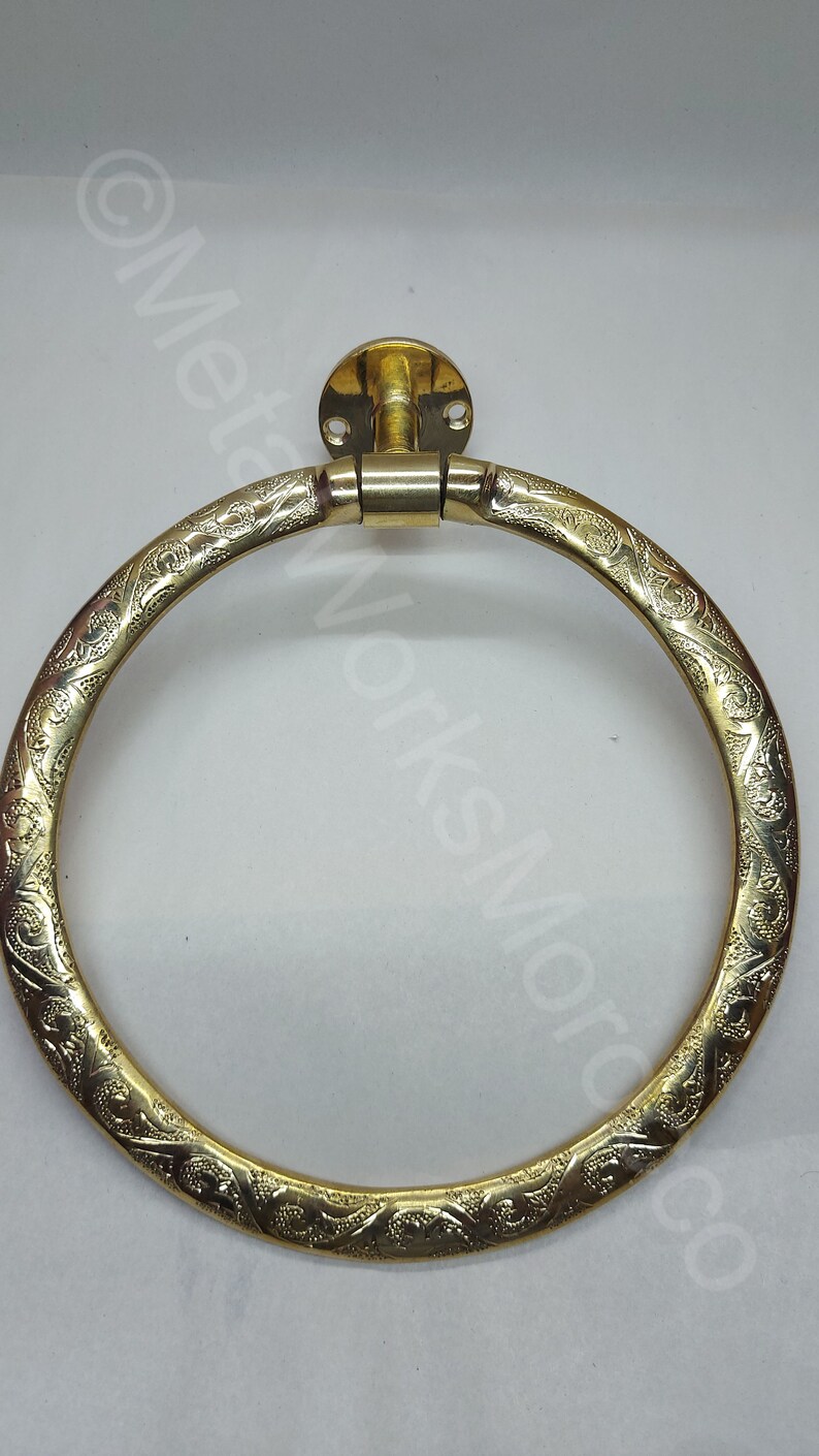 K&ouml;nnte beinhalten: Ein runder, dekorativer Handtuchring in Goldton mit einem komplizierten, gravierten Blumenmuster. Der Ring ist an einer Wandhalterung mit zwei Schrauben befestigt.