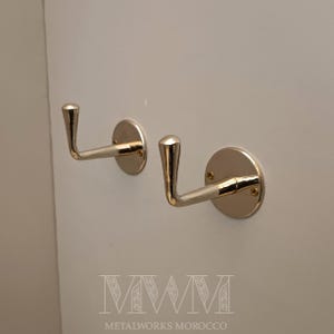 Puede incluir: Dos ganchos de pared de metal dorado con base redonda y gancho curvo. Los ganchos están montados en una pared blanca.